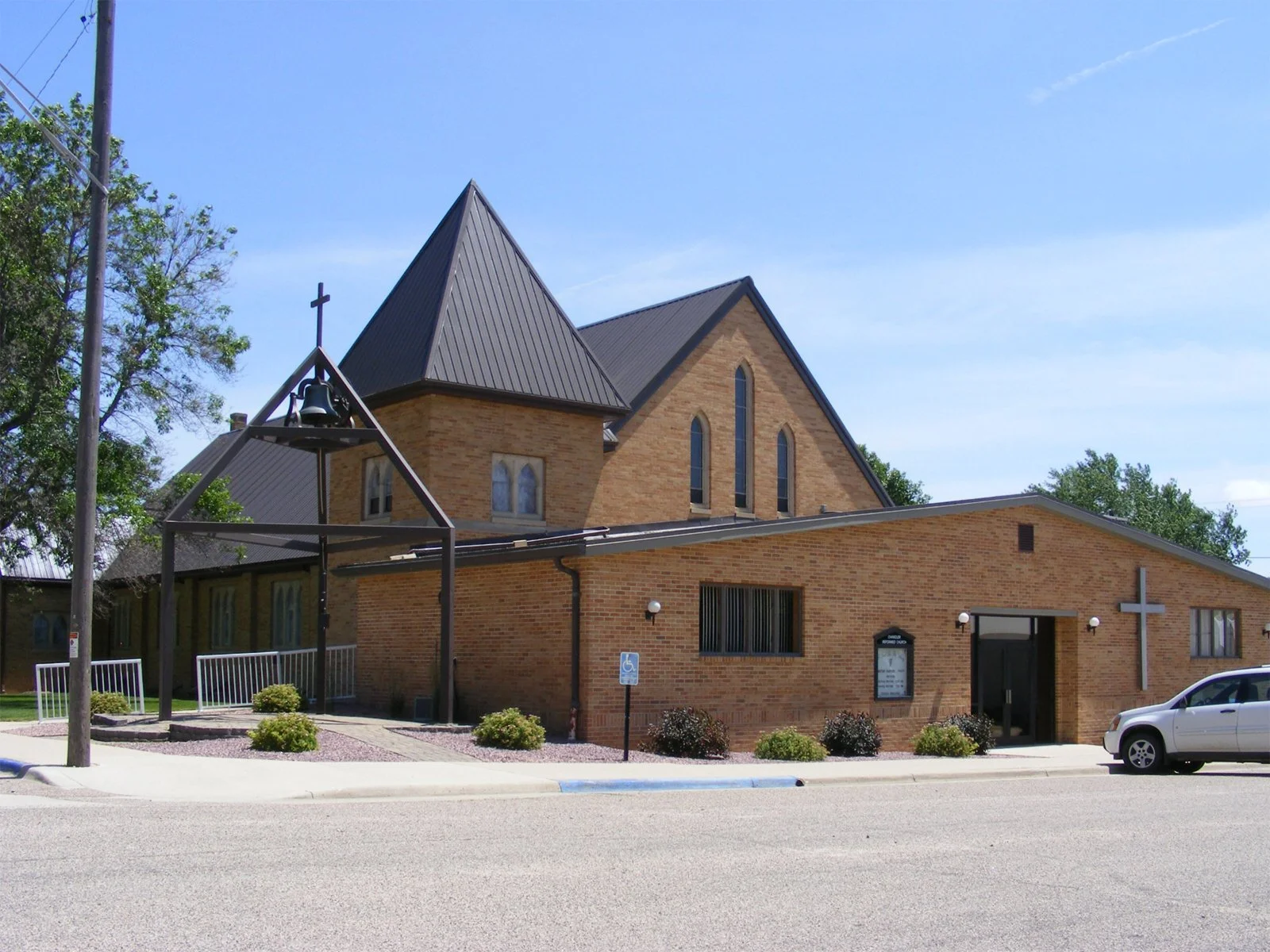 A brick church with a tall steeple topped with a cross, large windows, and a side entrance, located along a street with a parking lot and a silver car, under a clear blue sky.