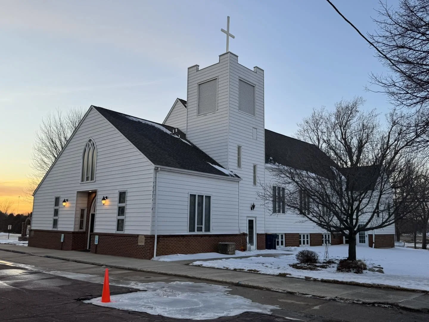 White wooden church with a steeple topped by a cross, set against a sunset sky, with snow on the ground and bare trees nearby.