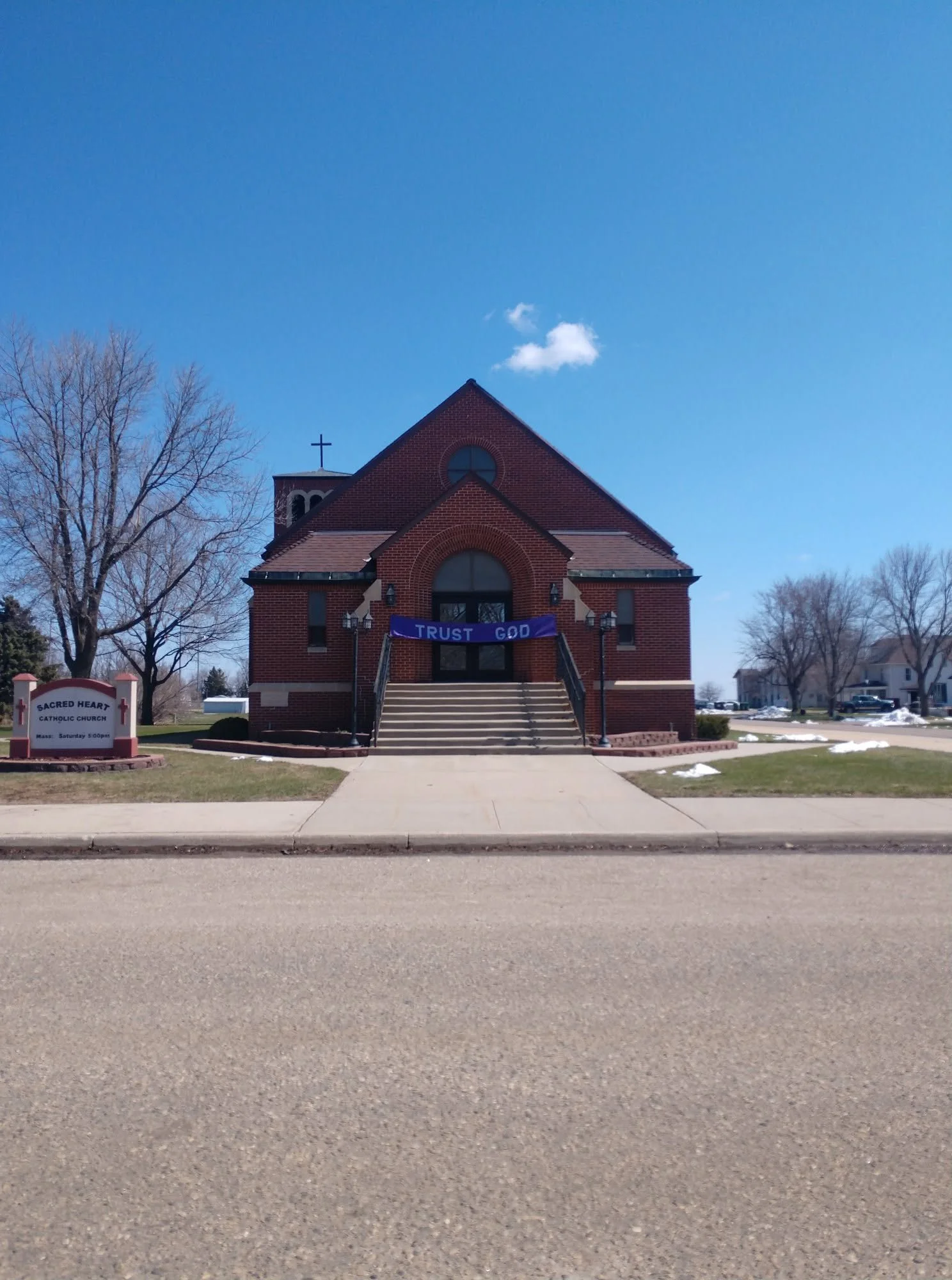 A brick church with steps leading up to the entrance, a purple banner with the words "Trust God," and a sign that reads "Sacred Heart Catholic Church". There are leafless trees and patches of snow on the ground under a clear blue sky.