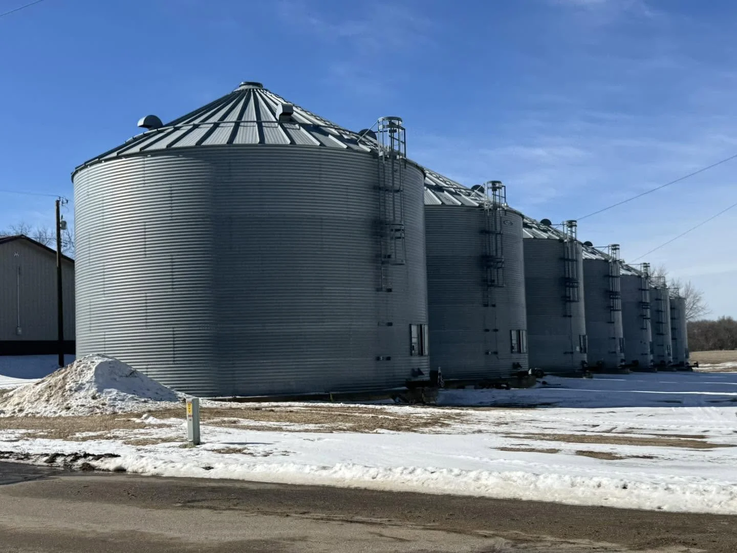 A row of large metal grain silos with rounded tops, situated outdoors on a snowy ground on a clear day with blue sky.