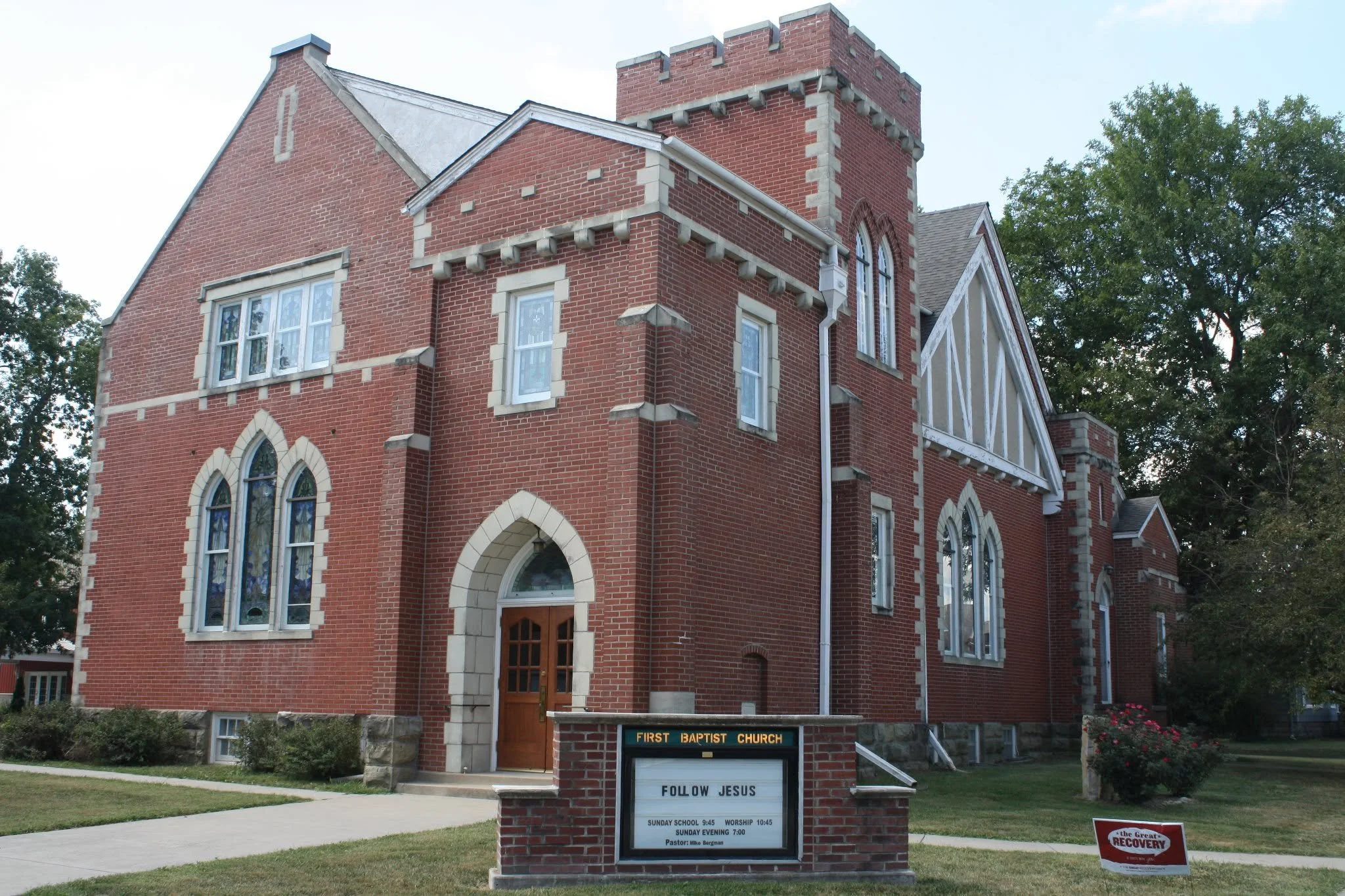 Brick church with a steeple and a signboard in front that reads 'First Baptist Church' and 'Follow Jesus'.