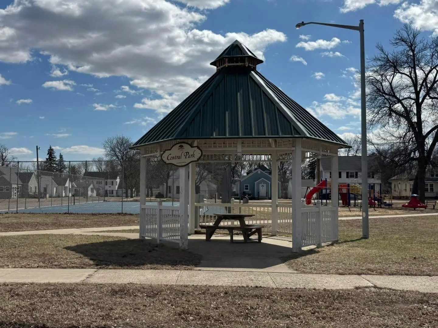 A park pavilion with a blue metal roof and a sign that reads 'Central Park'. There are playground equipment, a tennis court, and houses in the background. The sky is partly cloudy.