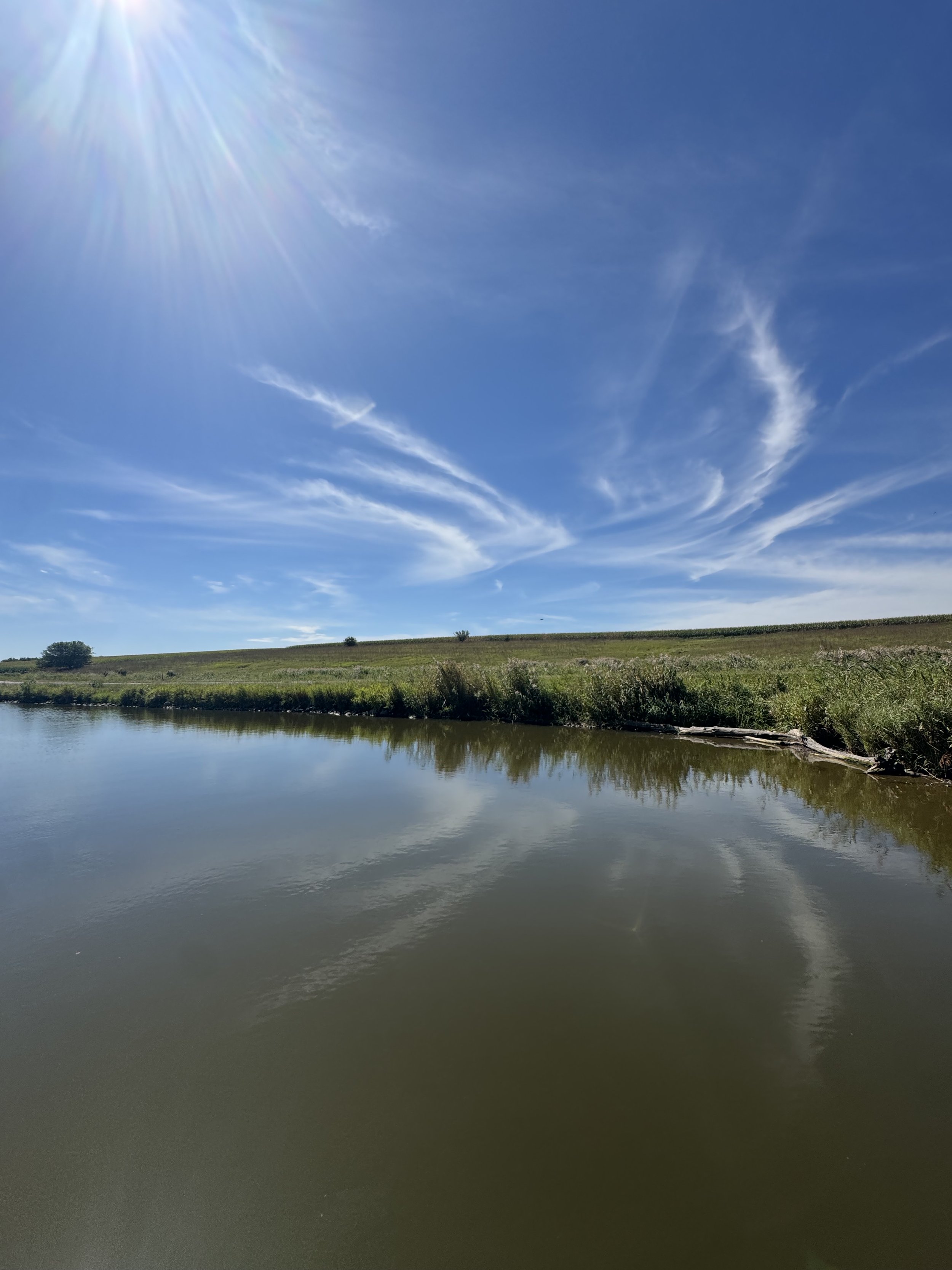 A peaceful landscape featuring a calm river with reflections of the sky, along with a grassy hill and a few small trees in the background, under a clear blue sky with wispy clouds and the sun shining brightly.