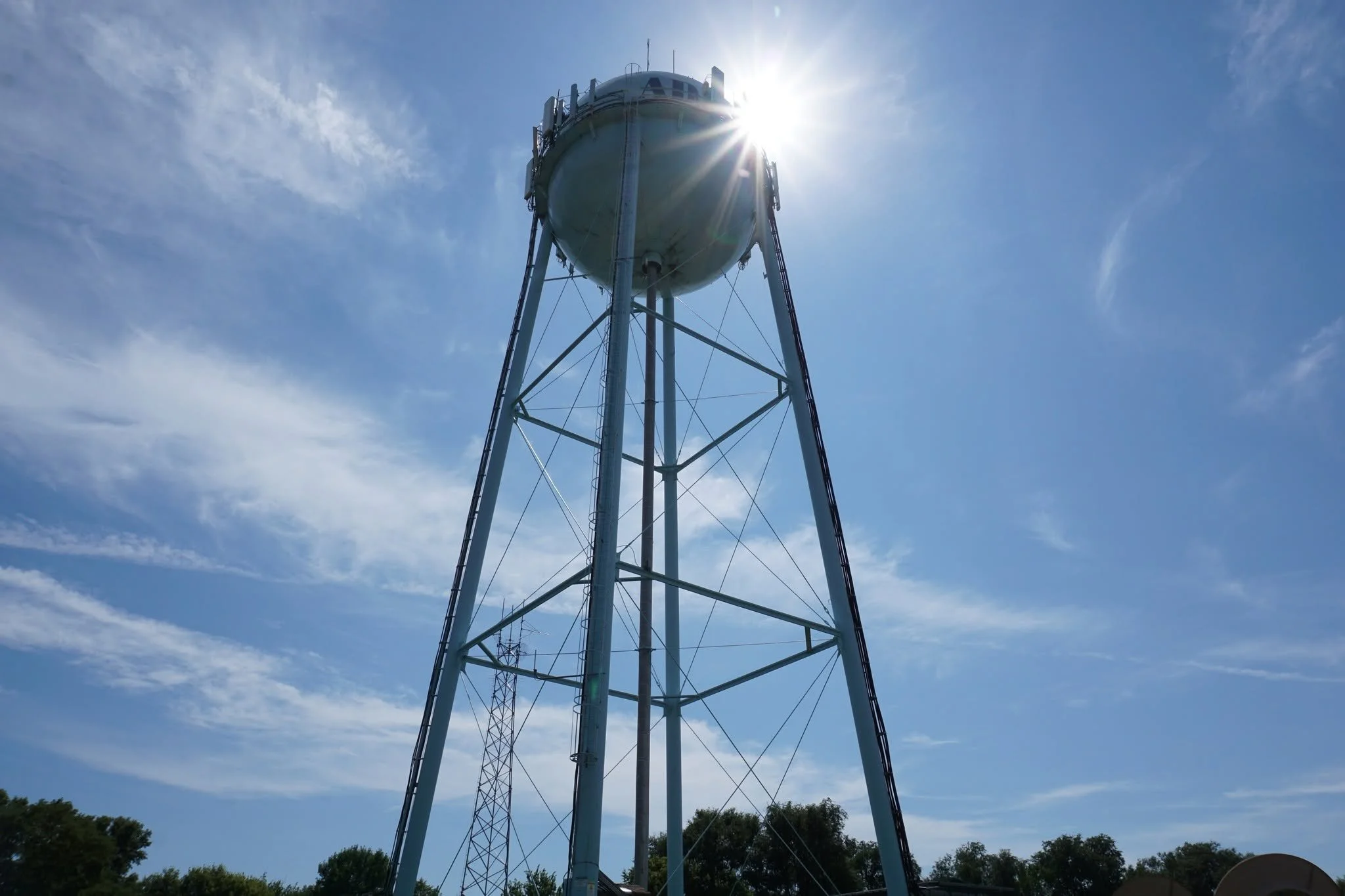 A tall water tower with a rounded tank at the top, silhouetted against a bright blue sky with some scattered clouds. The sun is shining from behind the tank, creating a lens flare effect.