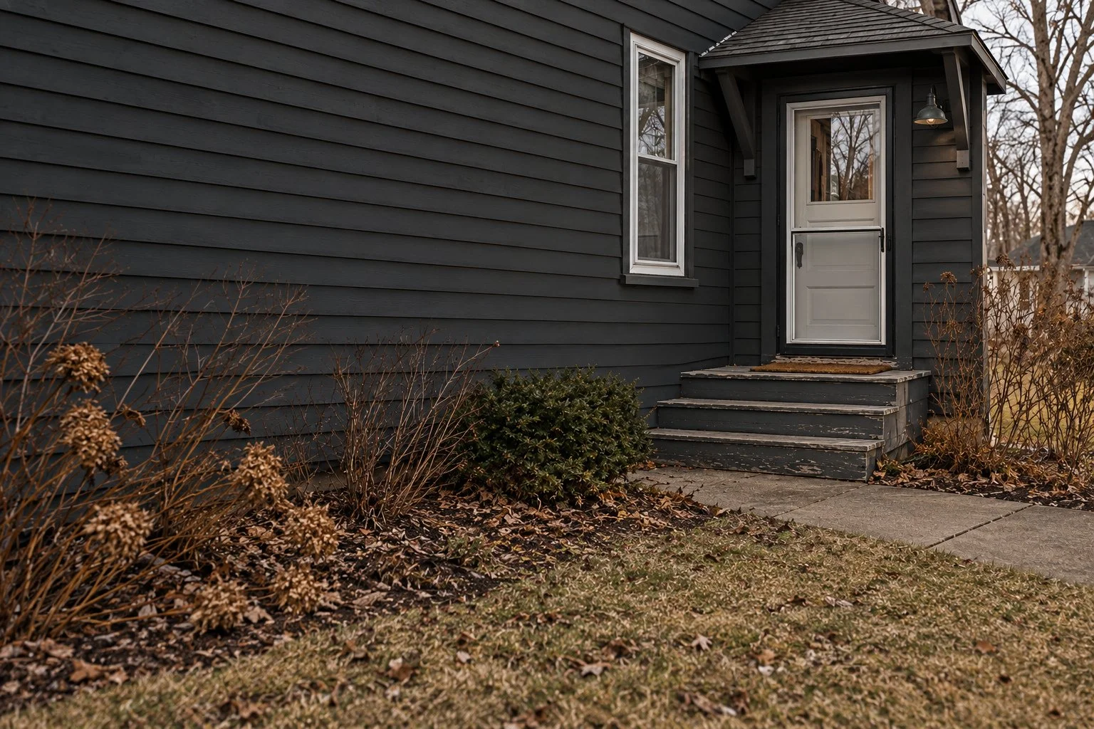 Front porch of a dark gray house with a white door, small set of steps, window to the left, and outdoor light fixture, with leafless bushes and grass in the yard.