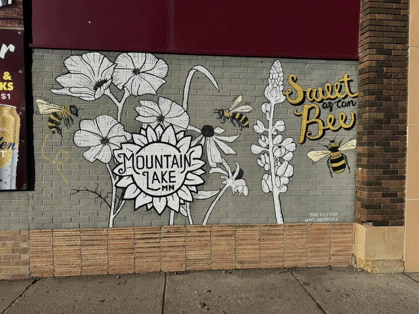 Street art mural on a brick wall featuring bees, flowers, and text that reads 'Mountain Lake MN' and 'Sweet as honey Bee' with yellow and black accents.