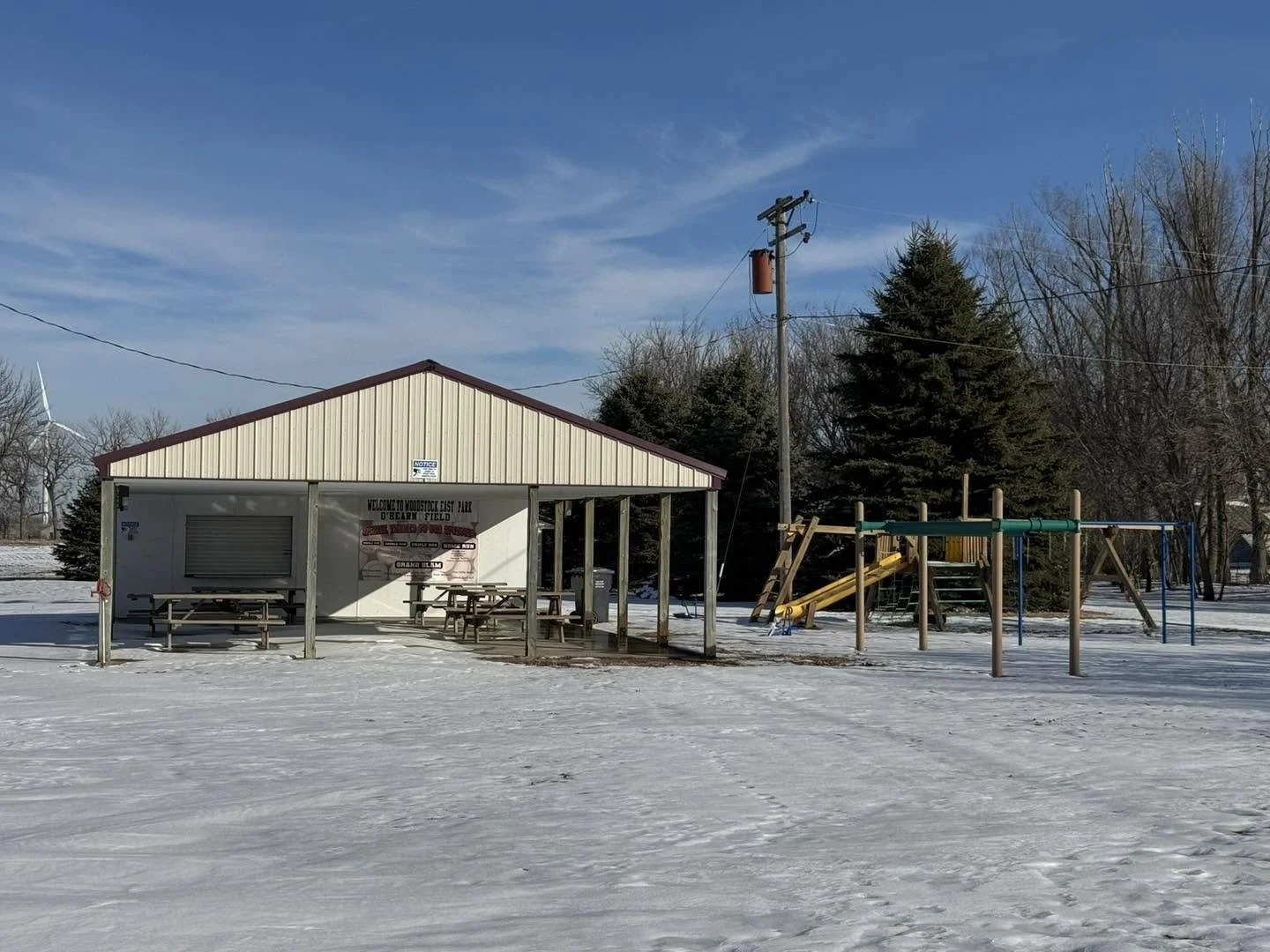 Snow-covered playground with a small pavilion, benches, and a set of swings and slides, near leafless trees on a clear day.