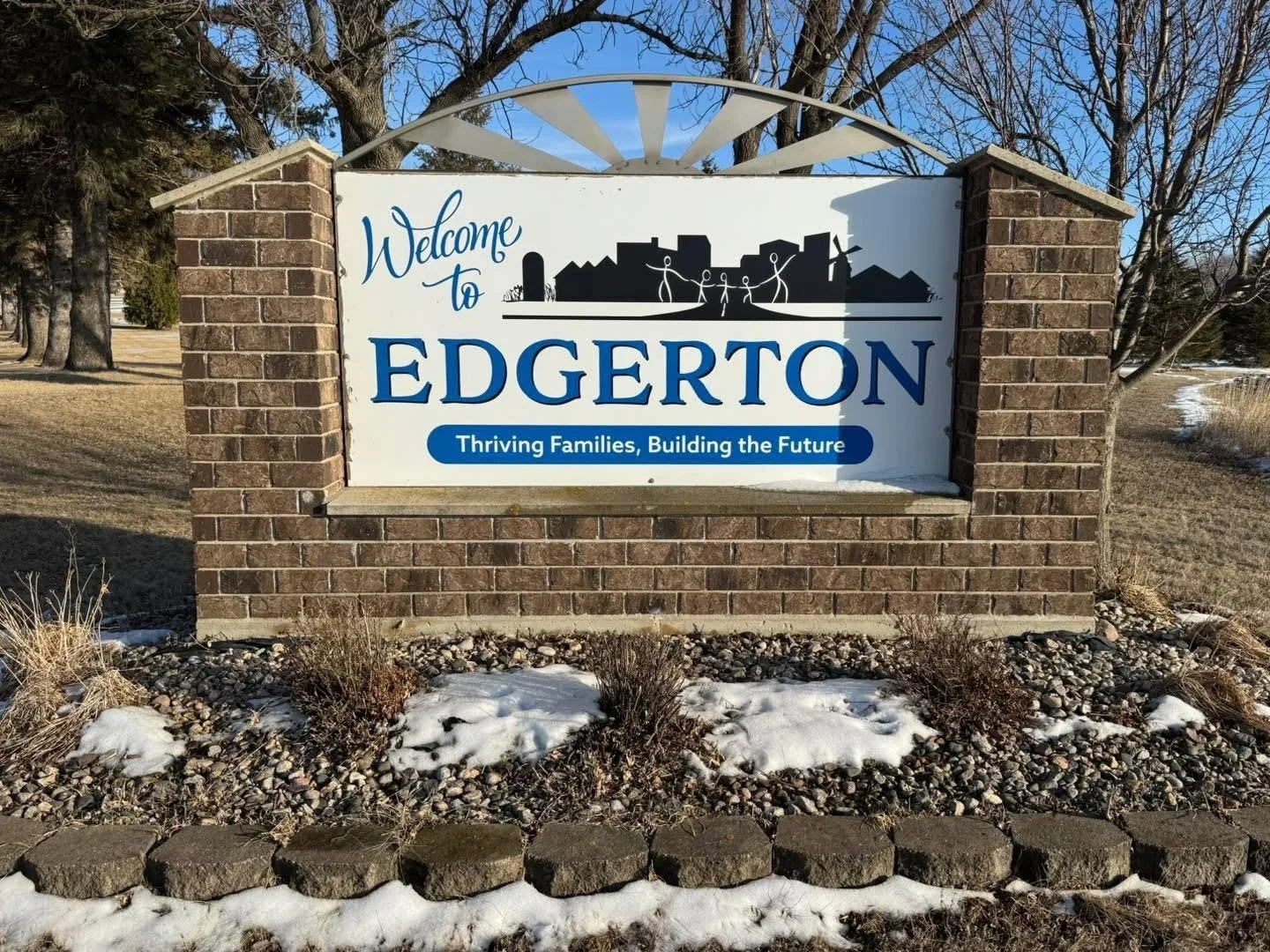 Welcome sign for Edgerton on a brick structure, with a cityscape silhouette and a phrase 'Thriving Families, Building the Future' on a blue banner, surrounded by trees and patches of snow.