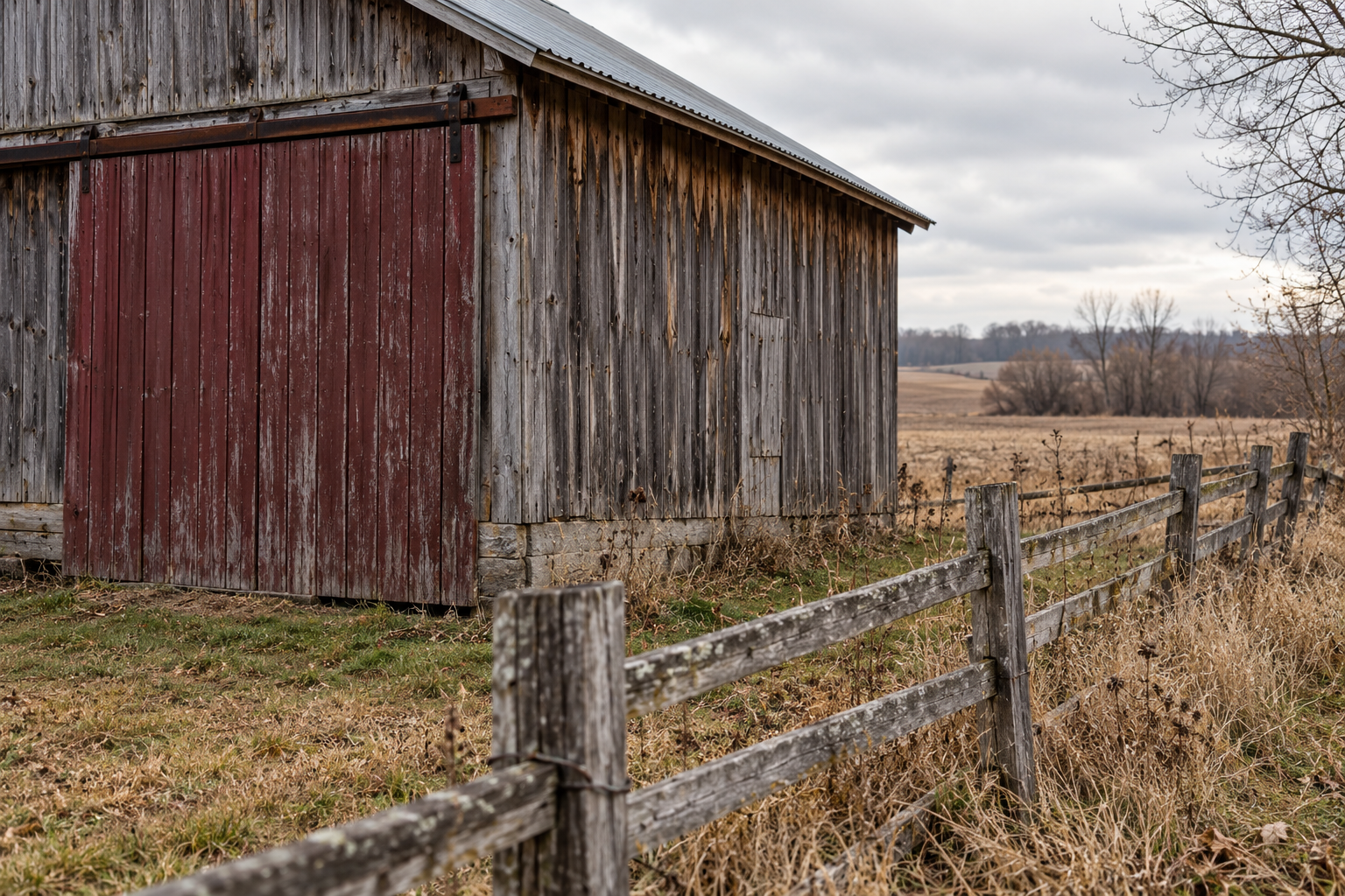 An old wooden barn on a rural landscape, with a weathered fence in the foreground, overcast sky, and distant trees and fields.