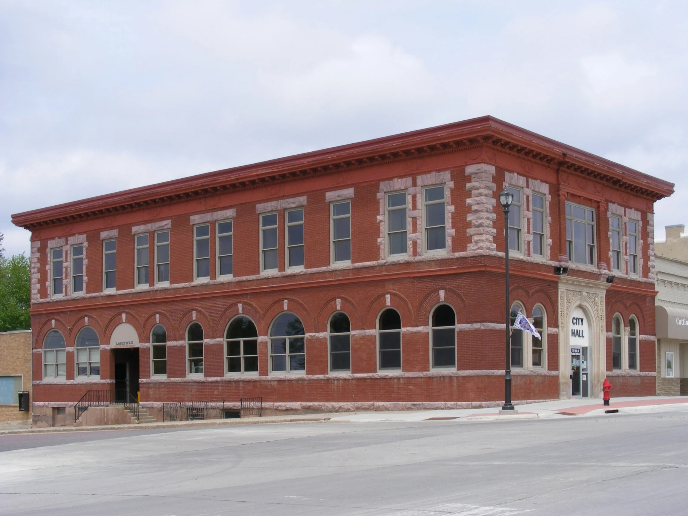 A red brick building with multiple windows and decorative stone accents, situated on a street with a lamp post and fire hydrant visible in the street corner, under a cloudy sky.