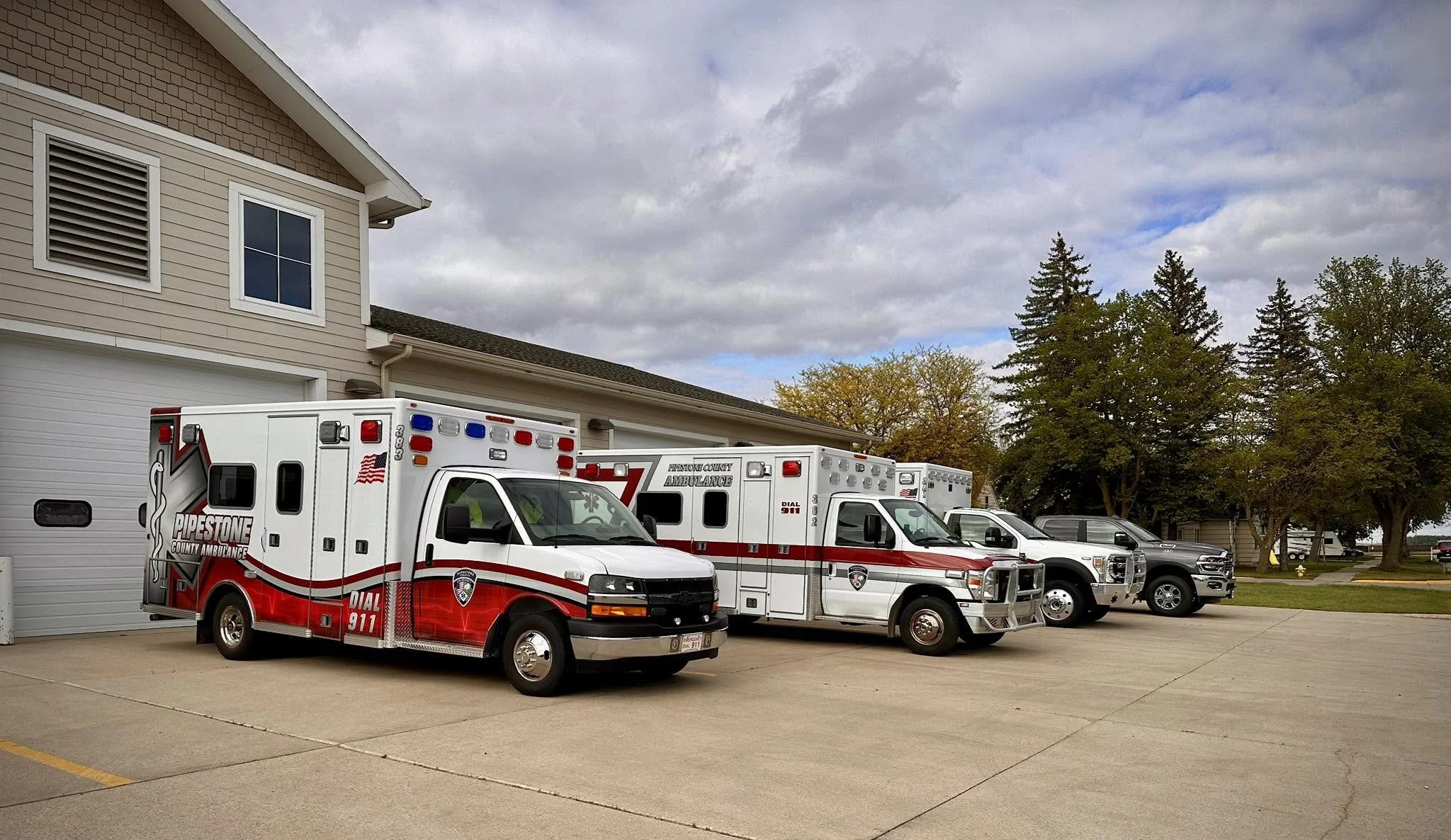 Two ambulances parked outside a residential building with a beige siding exterior and a white garage door, under a cloudy sky. There are also another vehicle and trees in the background.