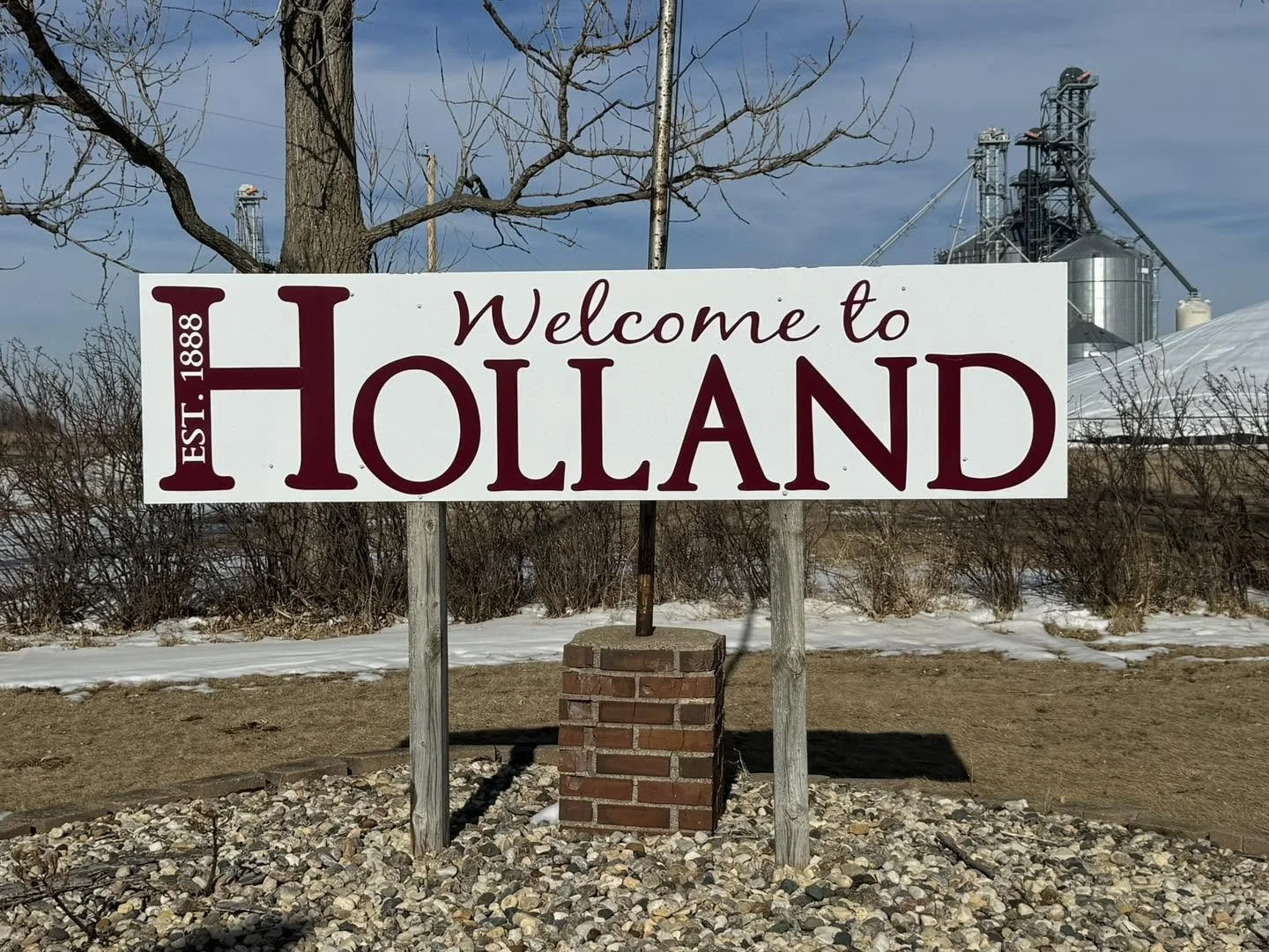 Welcome to Holland sign with establishment date 1888, placed on a brick pillar outside in winter with snow and leafless trees, industrial structures in the background.
