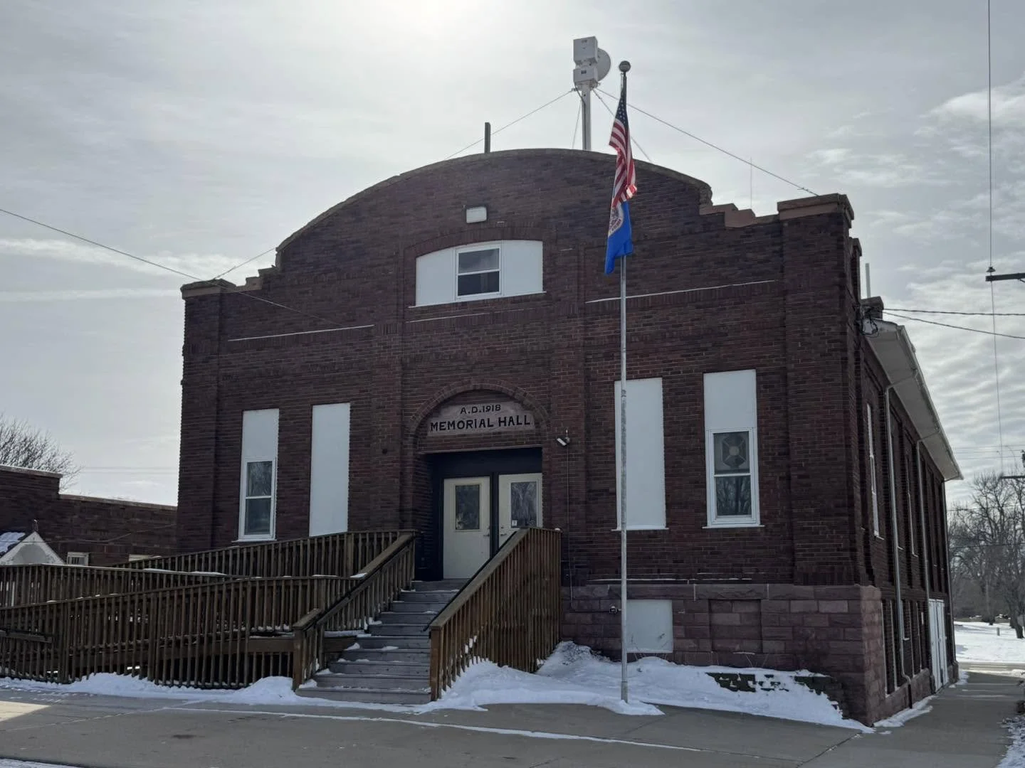 Red brick building labeled "Memorial Hall" with steps and wheelchair ramp, US flag and another flag on flagpole, snow on ground, cloudy sky.
