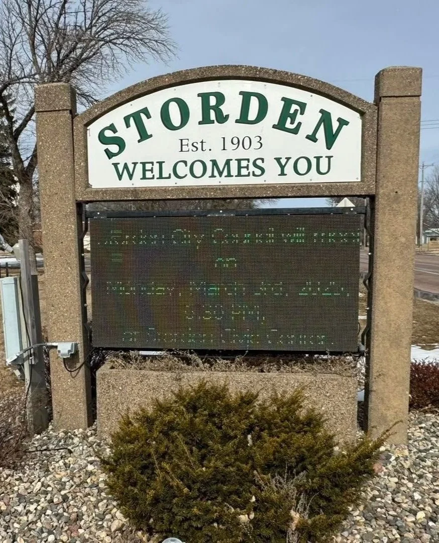 Sign for the town of Storden with the words "Established 1903 welcomes you" and an electronic message board below it, situated on gravel with a shrub in front.
