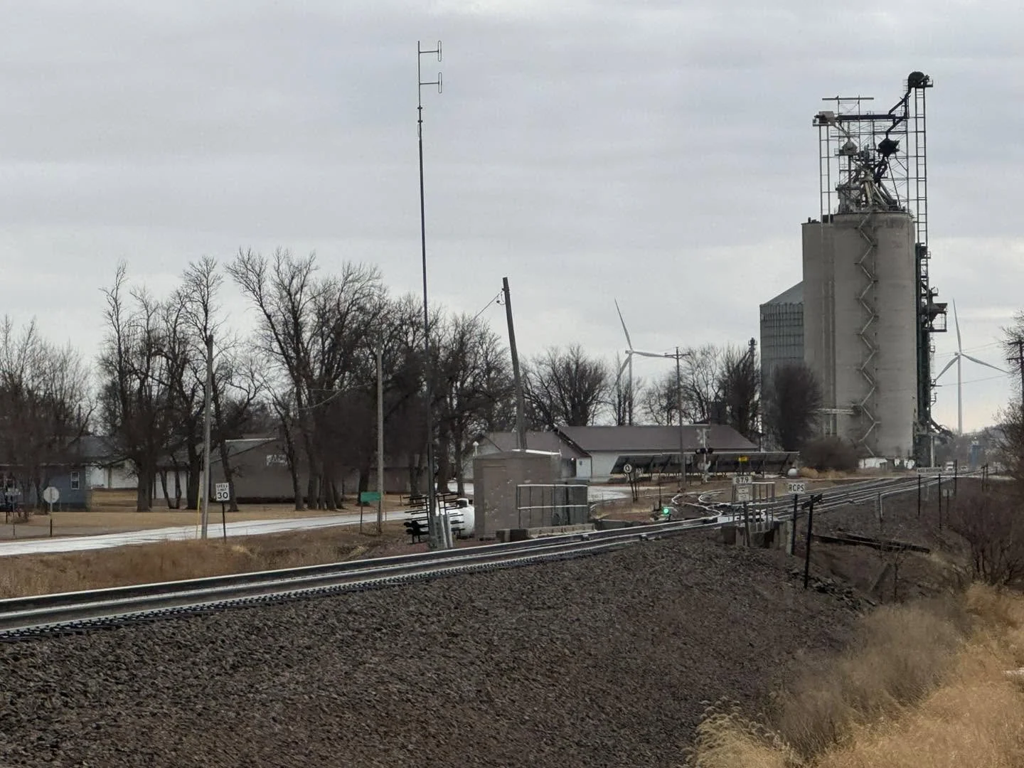 An industrial building with silos and wind turbines in a small town, viewed from a railway track with a signal light and utility poles, under a cloudy sky.