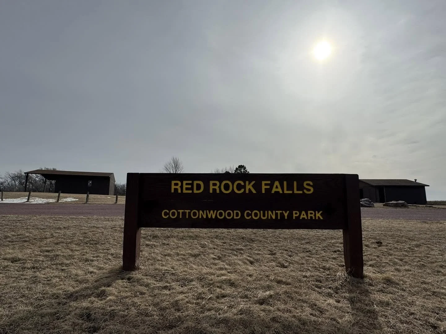 Sign for Red Rock Falls at Cottonwood County Park, with a grassy area, a building, and a clear sky with the sun.