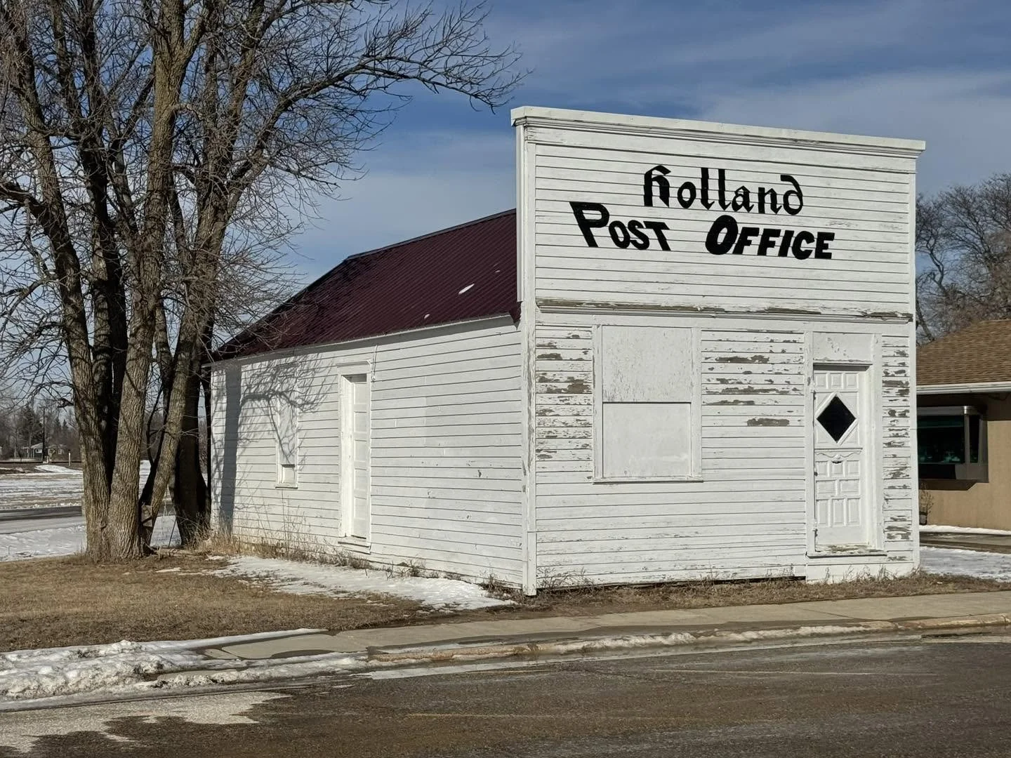 An old white wooden building with a sign that reads 'Holland Post Office' on the front. The building has a weathered appearance with paint peeling and boarded windows. There are trees nearby, some snow on the ground, and a sidewalk in front.