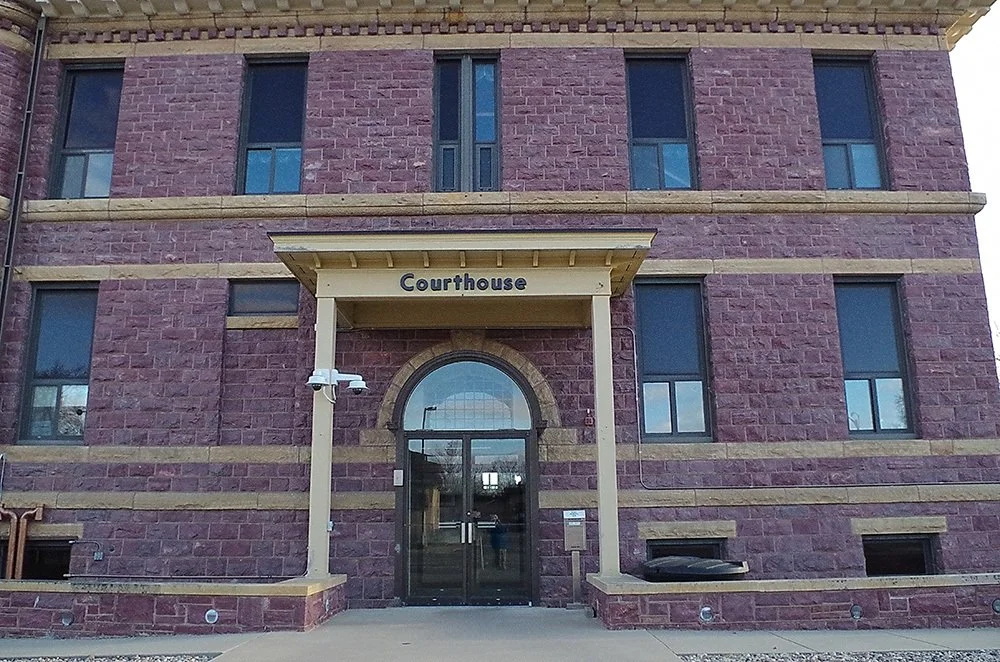 A three-story brick courthouse building with a sign above the entrance reading 'Courthouse'. The entrance has a glass door and an arched window above it, supported by two cream-colored columns. There are four windows visible on each floor, with some reflecting the sky. The building is made of red and brown bricks with tan horizontal bands and has small vent openings near the ground.