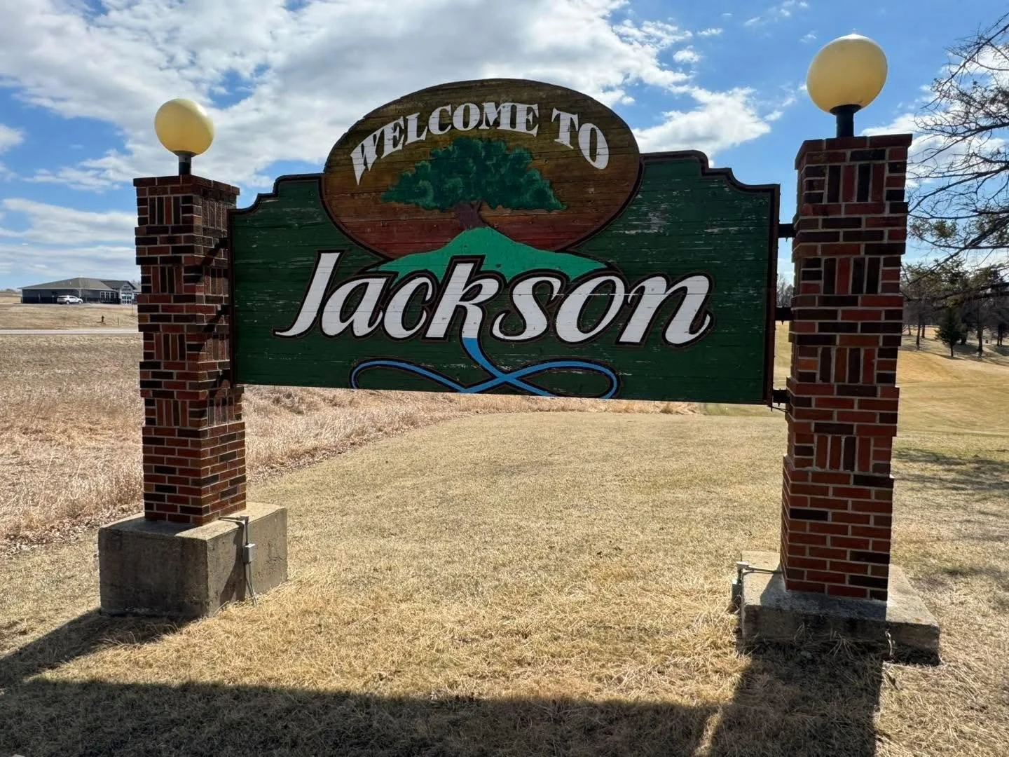 Welcome to Jackson sign with brick pillars and lamps, in a grassy area with a building in the background under a partly cloudy sky.