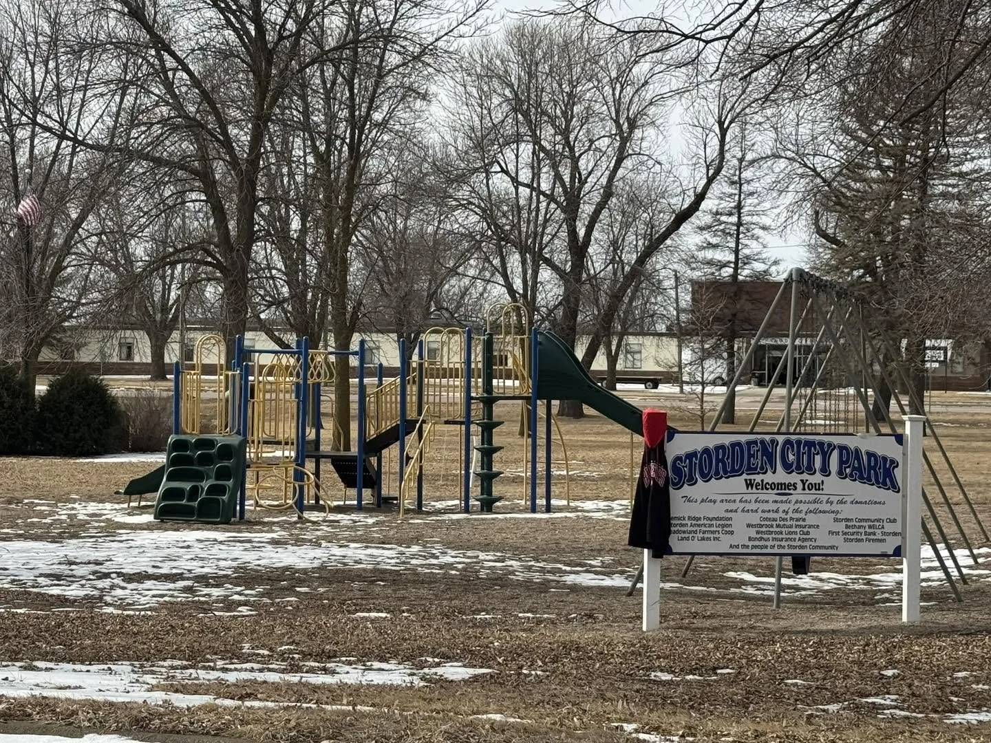Empty playground with a slide, swings, and climbing structures at Storden City Park on a cold day with patches of snow and leafless trees in the background.