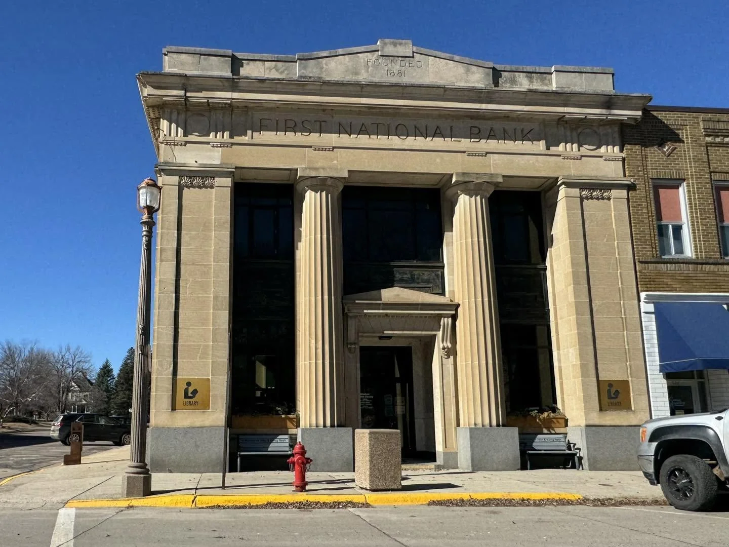 Historic building with columns, labeled 'First National Bank', with a sign indicating a library on the side, a fire hydrant, a parking meter, and a white truck parked on the street, under a clear blue sky.