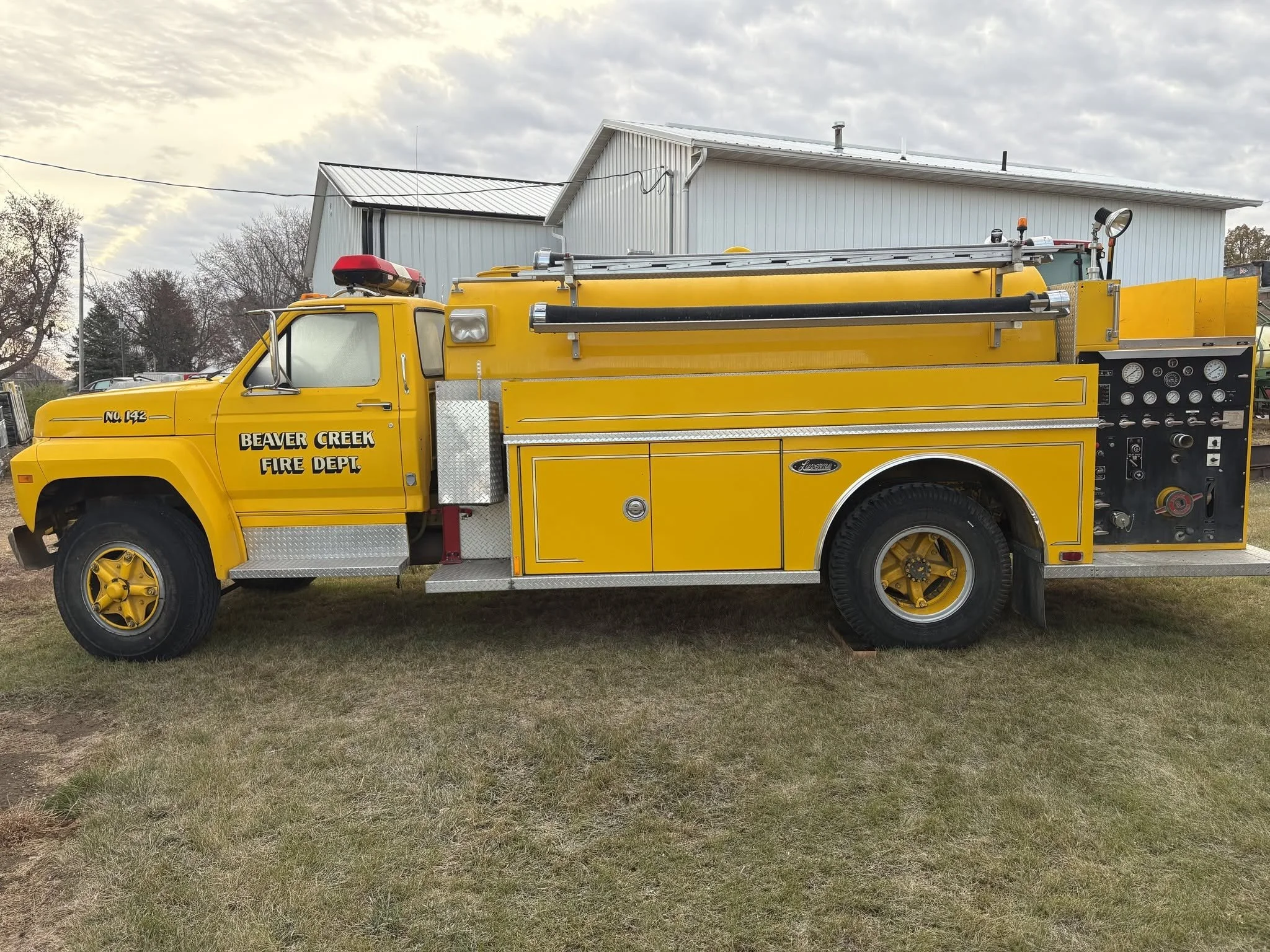 A yellow fire truck with 'Beaver Creek Fire Dept' on the door, parked on grass in front of a white industrial building with a metal roof, under a cloudy sky.