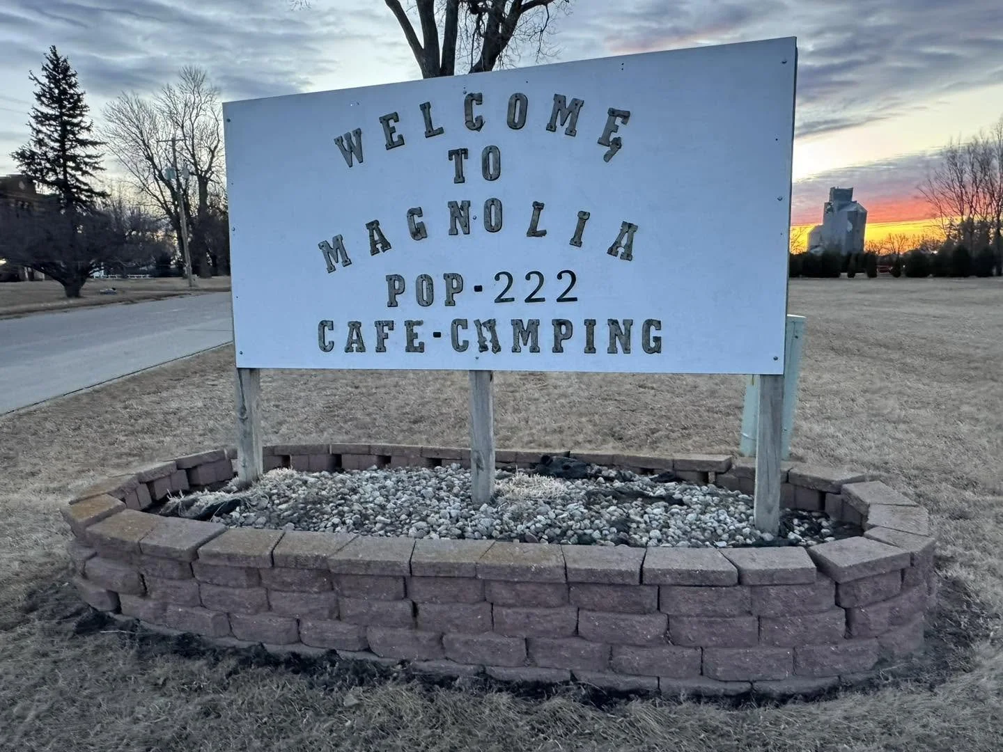 A large white welcome sign for the town of Magna, Utah, reading: 'Welcome to Magna, Pop. 222, Café, Camping'. The sign is mounted on wood with brick edging, situated in a grassy area near a road during sunset.