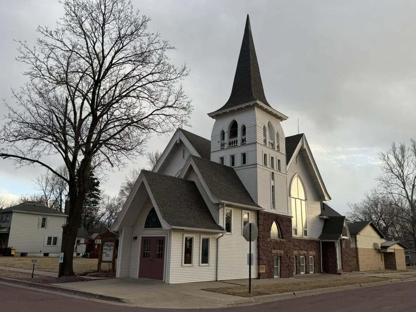 A white church with a tall steeple, surrounded by leafless trees, set against a cloudy sky.