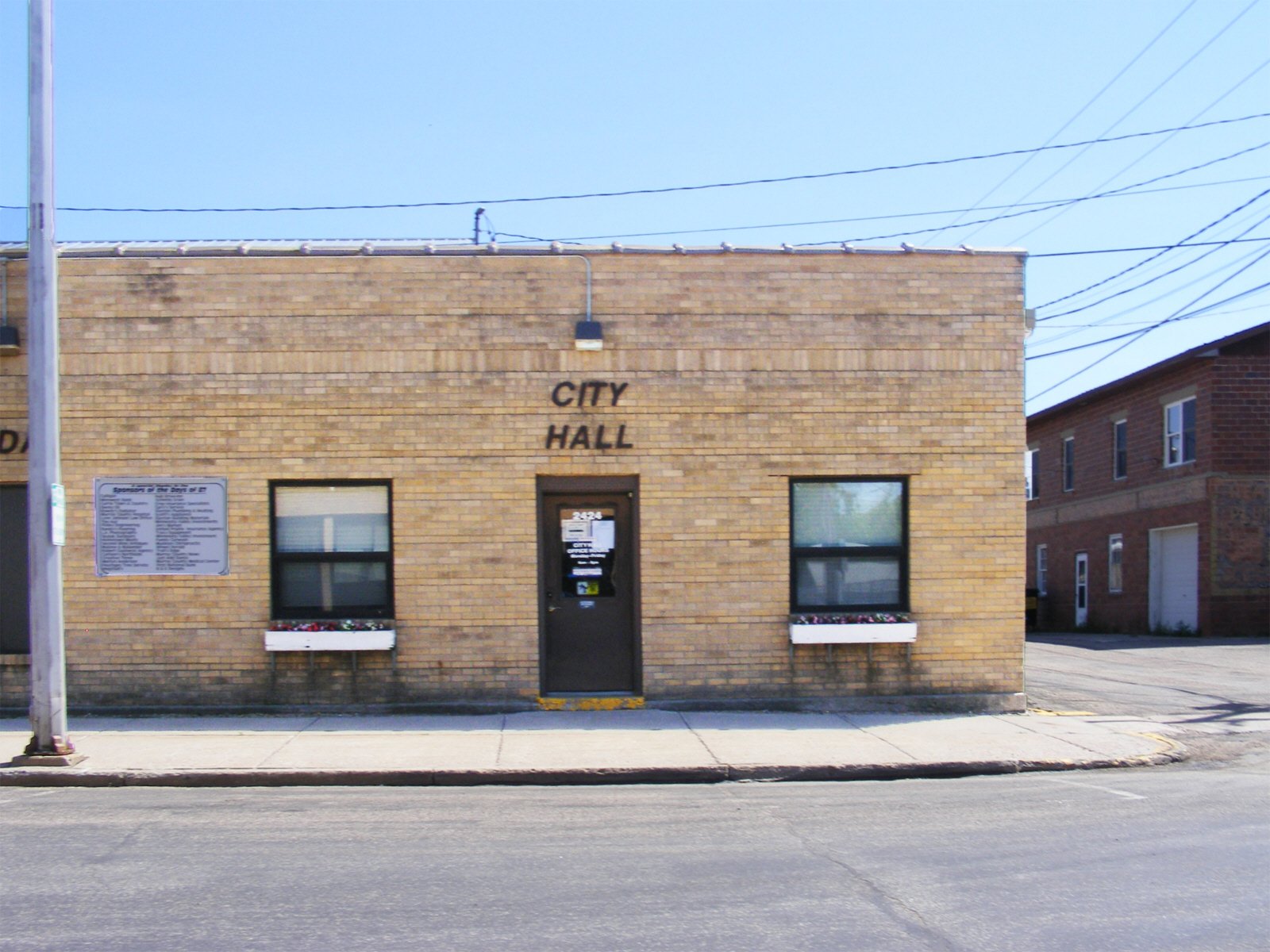 Brick city hall building with two windows with flower boxes and a black door in between, a sign on the left and a small plaque above the door, under a clear blue sky.