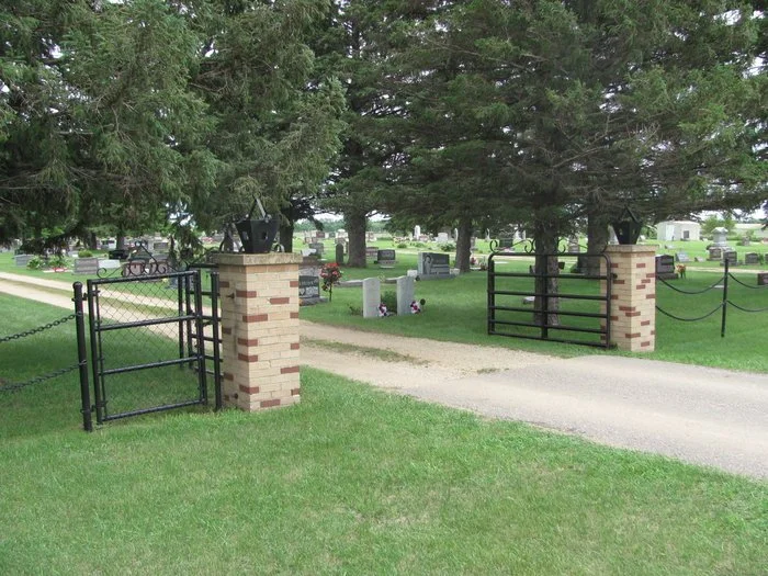 Entrance to a cemetery with cracked brick and black metal gate, gravel path, gravestones, and trees in the background.