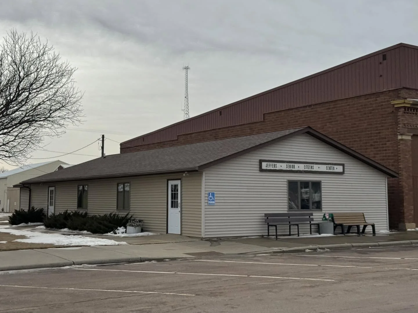 A small beige building labeled 'Jeffers Senior Citizens Center' with a bench outside, surrounded by a parking lot, with some snow on the ground, under a cloudy sky.