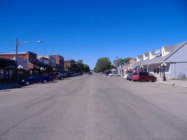 Main street in a small town with parked cars, storefronts, and a clear blue sky.