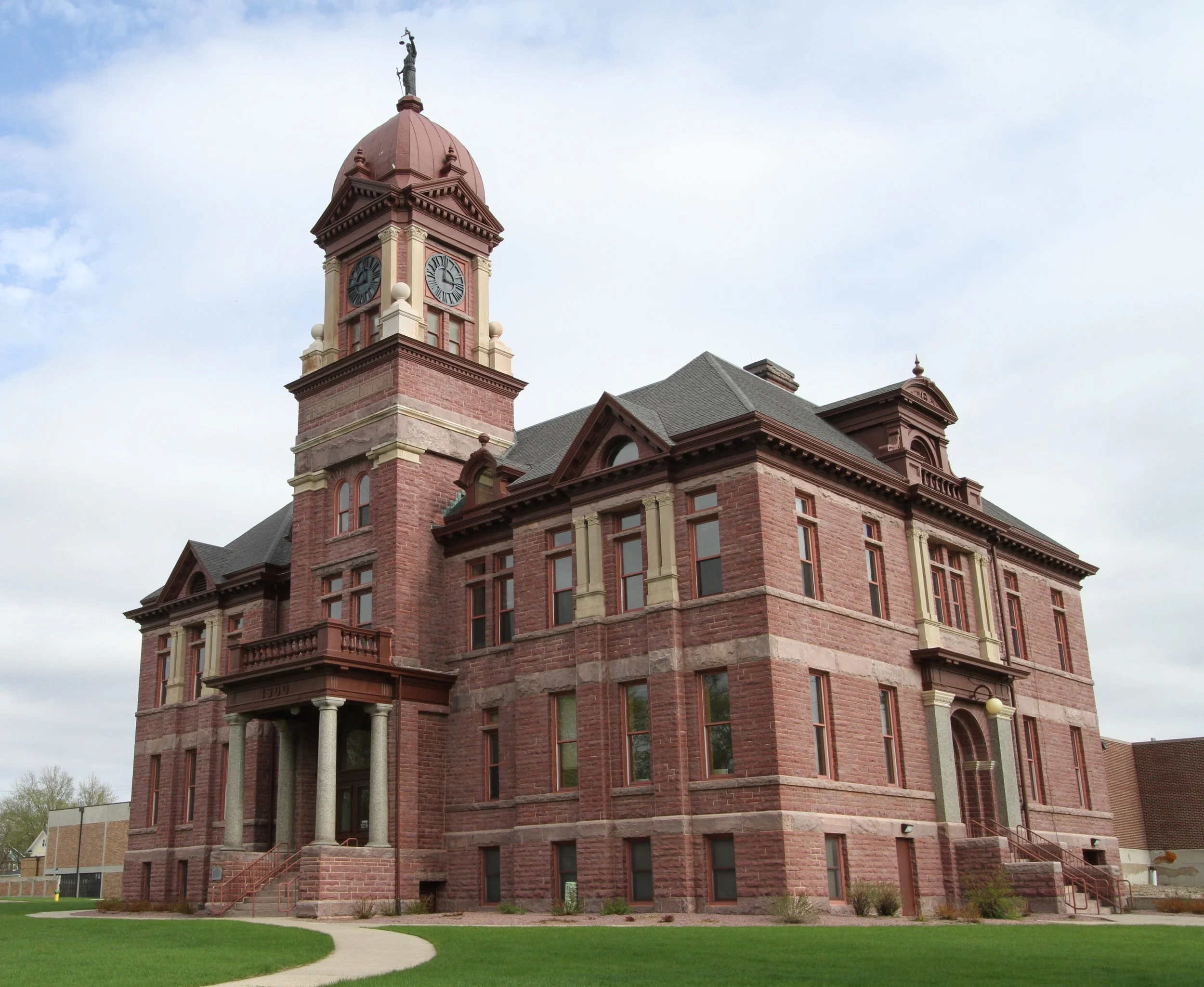 A historic red brick courthouse with a clock tower and statue on top, surrounded by a lawn with a walking path.
