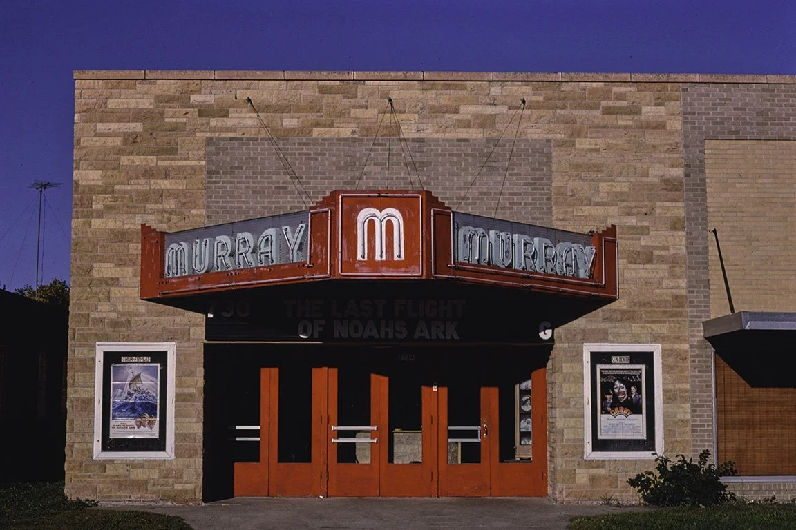The Murray Theatre with a vintage marquee sign displaying the name. Posters are on the brick wall beneath the sign, and the building has a tan brick exterior under a clear blue sky.