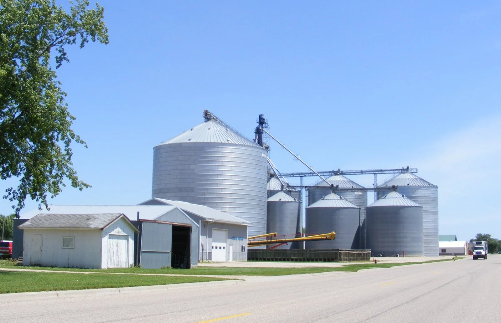 A farm with multiple metal grain silos and a small white storage shed under a clear blue sky.