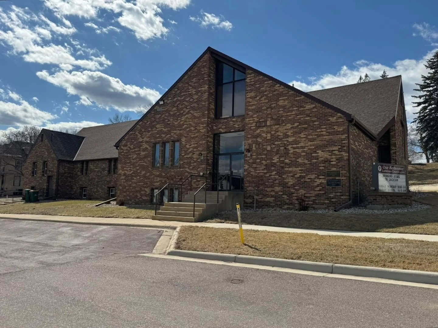 A brick church building with a front staircase, small grassy area, a sidewalk, a parking lot, and a blue sky with clouds.