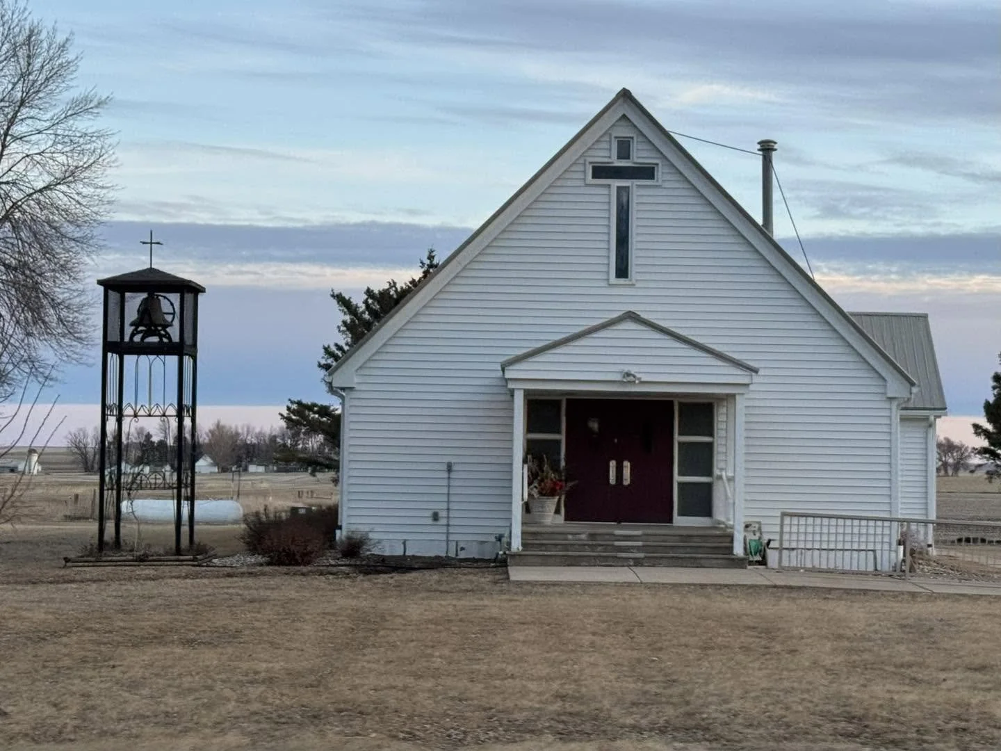 A white church with a steeple and a large cross on the front, situated in a rural area with open fields and a cloudy sky in the background.