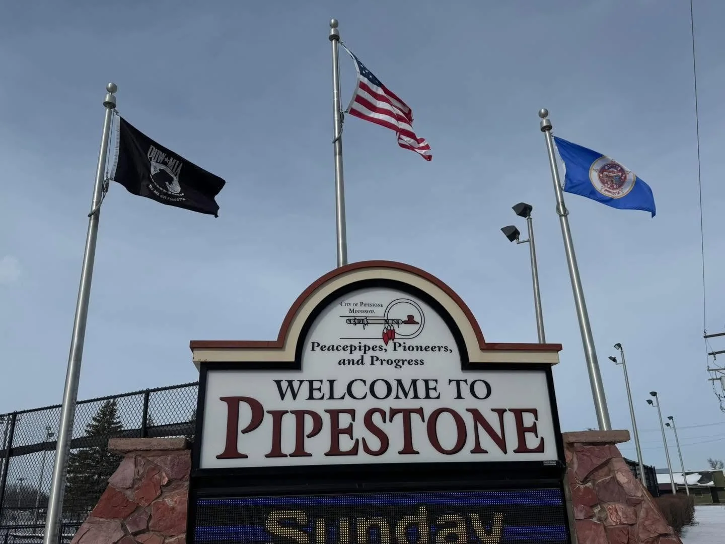 A welcome sign for Pipestone, Minnesota, with three flags flying above it and a cloudy sky in the background.