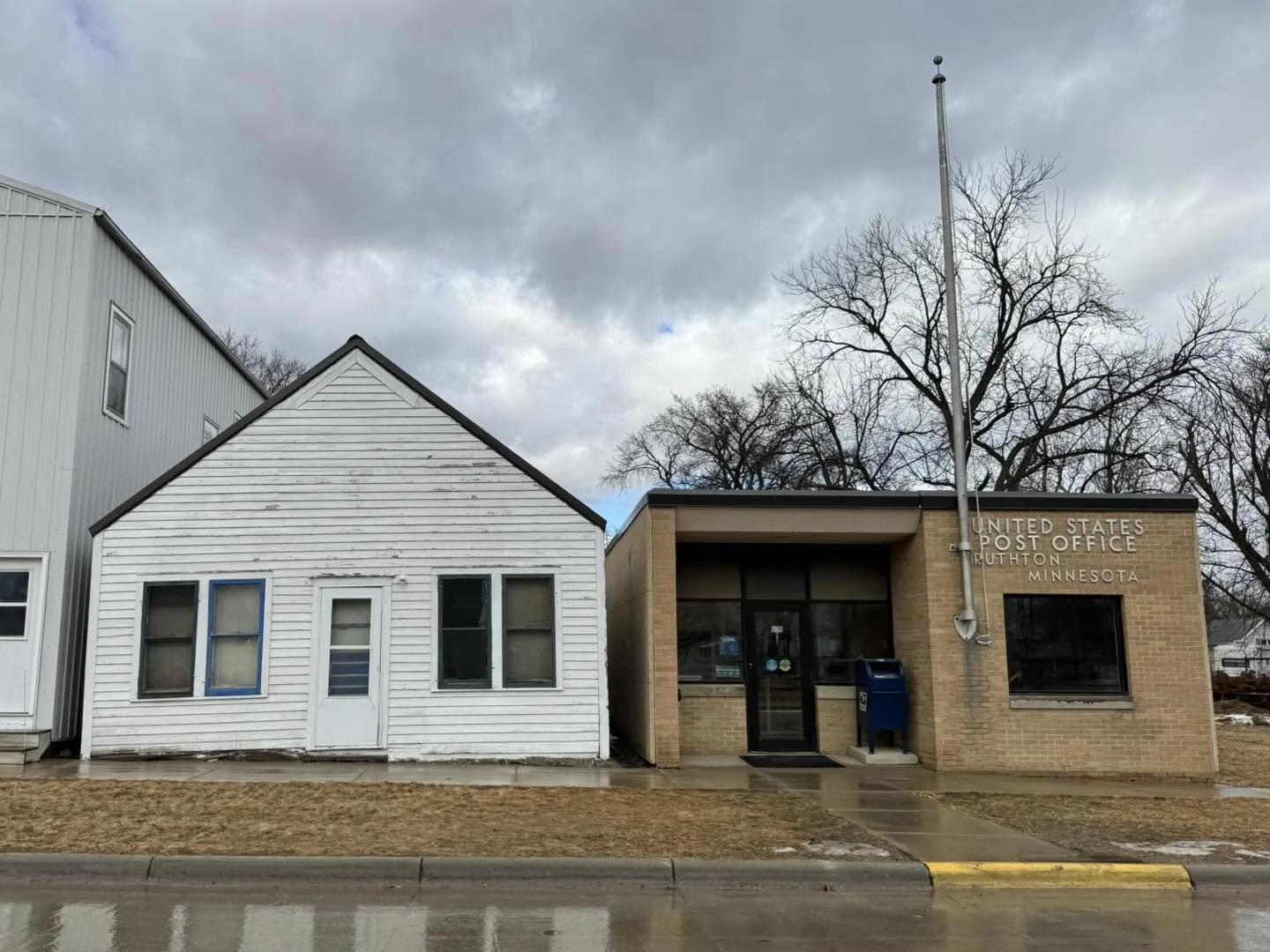 The image shows a small white wooden building with three windows and a door, next to a modern brick post office with a sign reading "United States Post Office RUTHON MINNESOTA." There are leafless trees behind the buildings and a cloudy sky above.