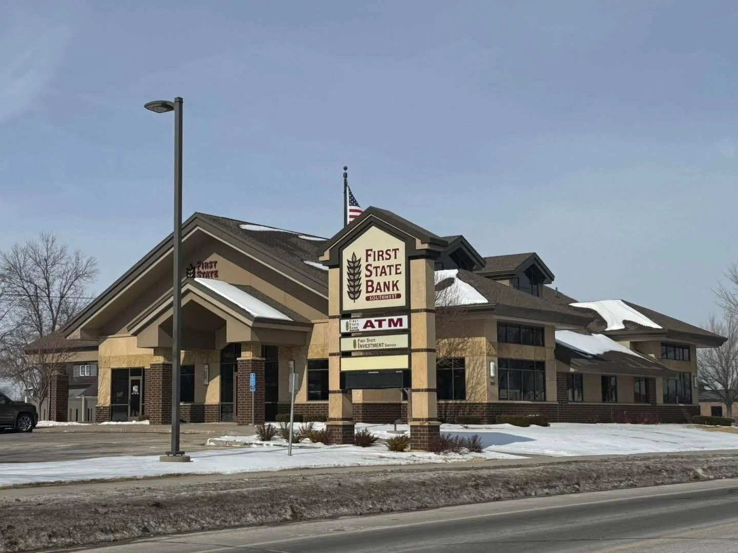 A brick and beige commercial building with a sign reading First State Bank, featuring an ATM, under a partly cloudy sky with snow on the ground.