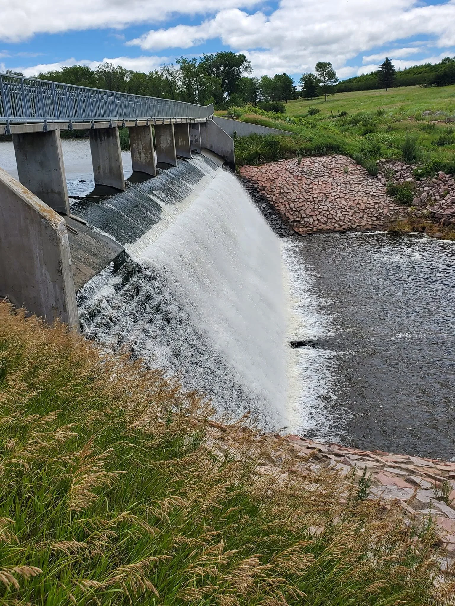 A dam with water spilling over, surrounded by green grass and trees, under a partly cloudy sky.