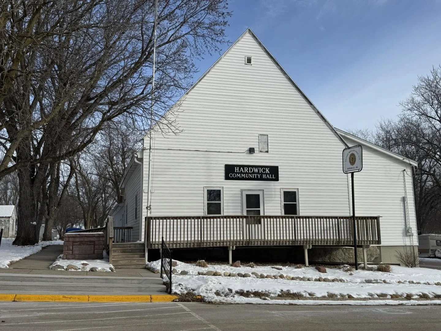 White wooden community hall building with black sign that reads 'Hardwick Community Hall' in a snowy landscape with leafless trees and a sidewalk in the foreground.