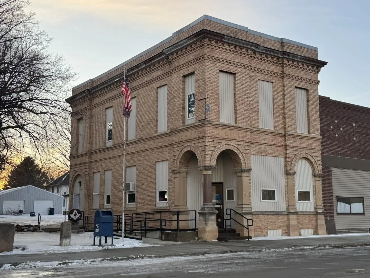 Historic brick building with flags, a U.S. flag and a Masonic flag, and a mailbox in front. Snow on the ground, late afternoon or early evening.