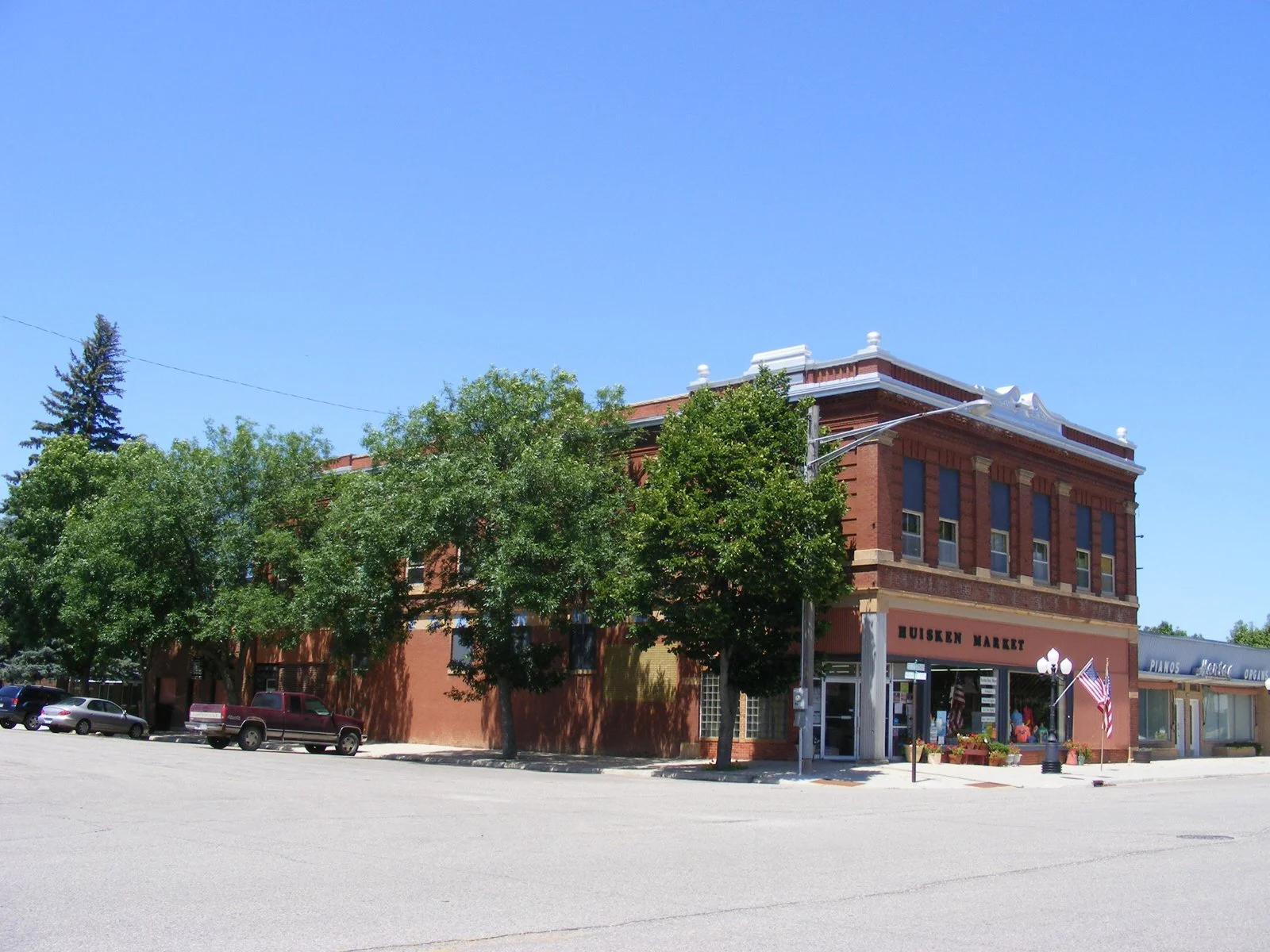 A historic brick building with a sign reading Liusken Market on the front, surrounded by trees, parked cars, and a clear blue sky.