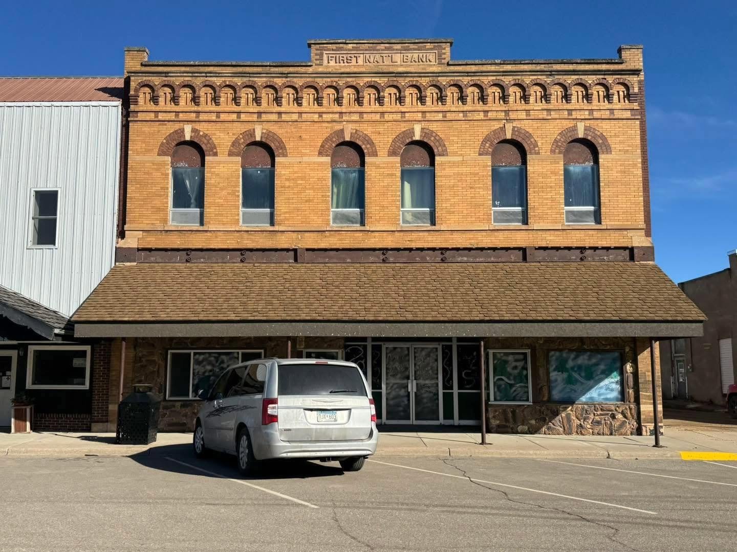 A two-story brick building with a sign that reads "First Natl Bank" on the upper facade. The building has five tall arched windows on the second floor and a large glass door on the ground floor. There is a gray minivan parked in front of the building, along with a parking lot and a small trash bin. The sky is clear and blue.