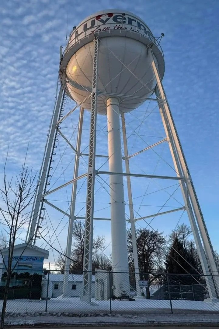 A tall white water tower with a spherical tank at the top, featuring lettering and artwork, enclosed by a chain-link fence, with snow on the ground and leafless trees underneath a partly cloudy sky.