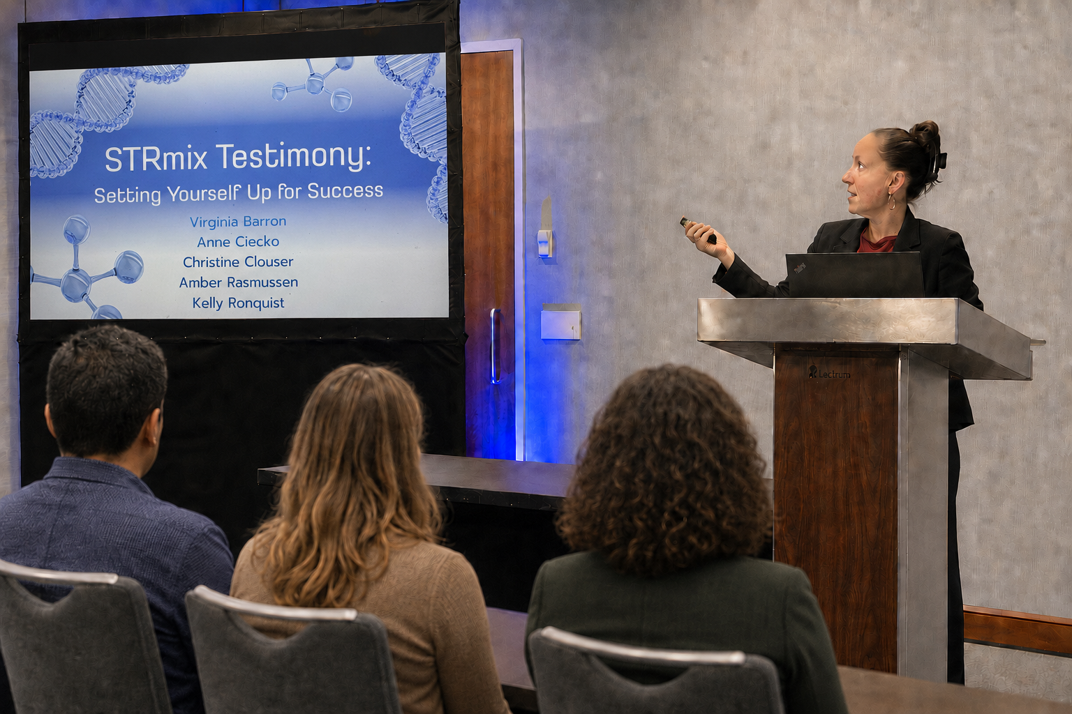 A woman giving a presentation at a conference, standing behind a podium with a laptop, pointing towards a large screen displaying a slide titled 'STRmix Testimony: Setting Yourself Up for Success' with names listed. Three audience members are seated and listening.
