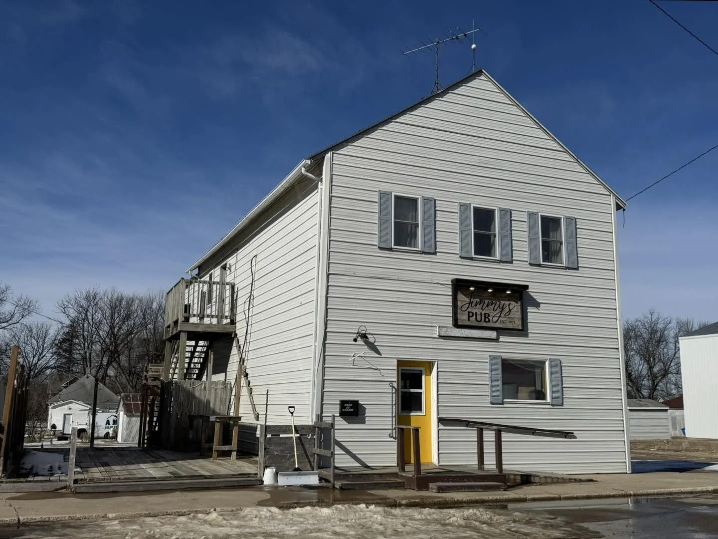 Two-story white building with blue shutters, yellow door, sign reading 'Jimmy's PUB' on the front, and outdoor staircase.