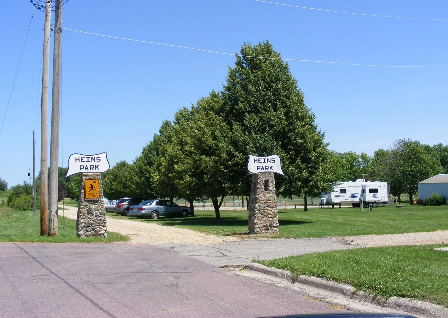 Entrance to Heins Park with two stone pillars and signs that read 'Heins Park' and a small yellow sign that says 'Slow children at play'. There is a gravel driveway with parked cars, a grassy area with trees, a white RV trailer, and some buildings in the background under a clear blue sky.