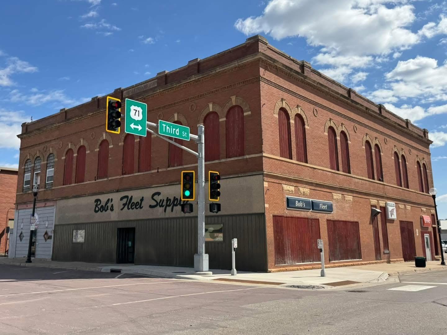 A two-story brick building at the corner of an intersection, with a sign reading 'Bob's Fleet Supplies' on the front, traffic lights, street signs for Third Street and Route 71, and a mostly clear sky with a few clouds.